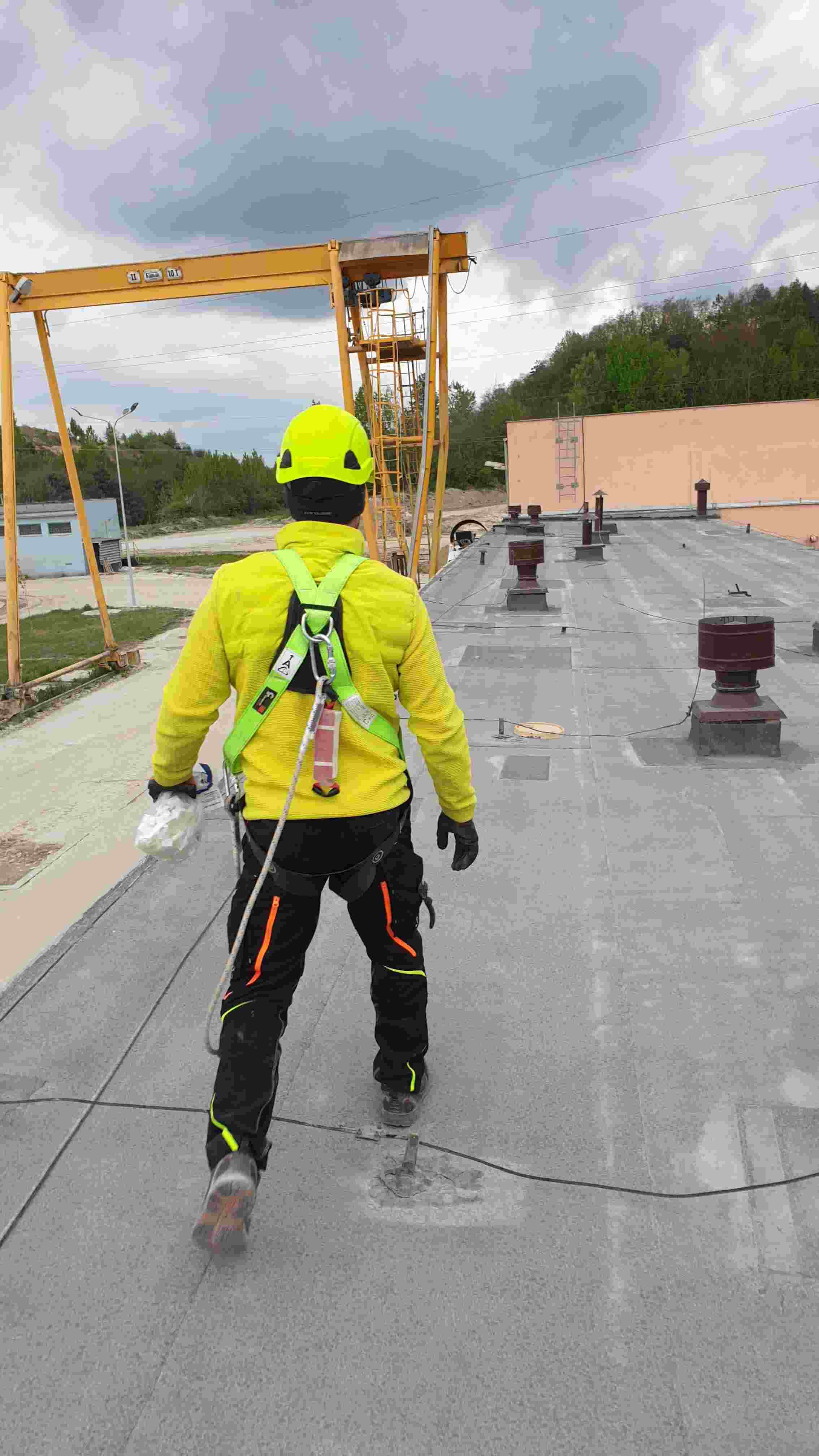 Technik w żółtej kurtce na dachu hali – prace instalacyjne z szelkami